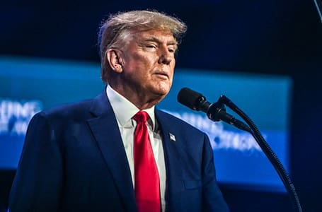 Donald Trump standing behind a podium speaking to a crowd. He's in a blue suit with a red tie.