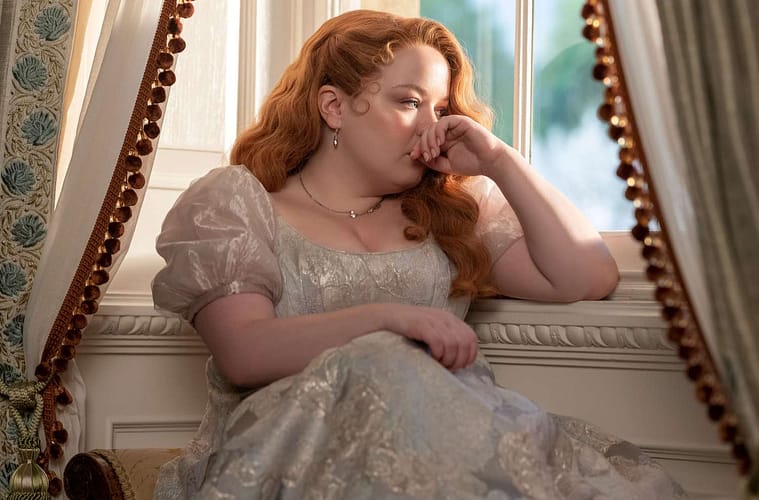 Penelope Featherington sitting by a window staring longingly outside with her hair down and wearing 'Bridgerton Blue'.