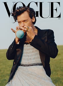 Harry Styles on the cover of Vogue Magazine wearing a baby blue lace dress with black trimming and a black blazer on top. He's blowing into a small baby blue balloon and is set on a field of green grass.