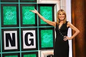Vanna White gestures at a wheel of fortune puzzle in a black dress