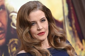 Lisa Marie Presley posing on a red carpet wearing an off the shoulder navy dress with red lipstick and loosely curled hair.