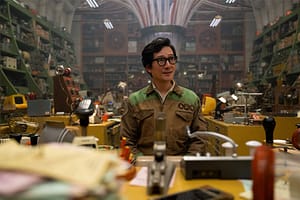 Ke Huy Quan as Q.B in Loki season 2. His is wearing glasses, a TVA uniform and looks to be in a large workshop.