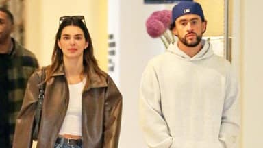 Kendall Jenner and Bad Bunny walking together. Bad Bunny is in a grey sweatshirt and a blue hat. Kendall is wearing a white crop top with a large light brown leather jacket, blue jeans with a black belt and a gold clasp, black sunglasses on her hair and a black handbag on her shoulder.