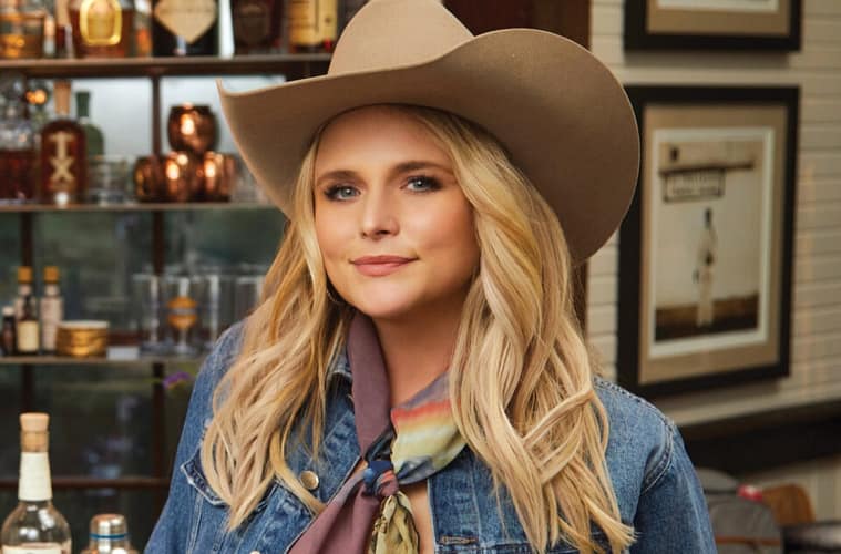 Miranda Lambert sitting at a bar posing towards the camera with a slight grin. she is wearing a jean jacket and a light tan cowboy hat.