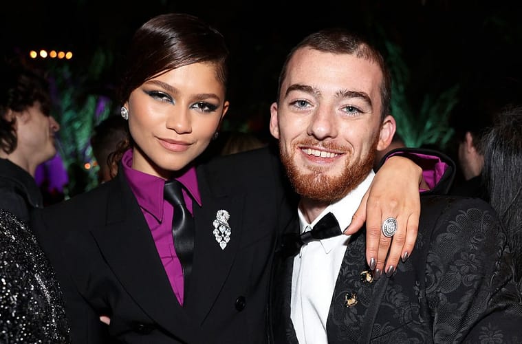 Zendaya (left) in a black suit and tie with a vibrant purple shirt, with her arm around Angus Cloud (right) who's wearing a black tux with baroque detailing.