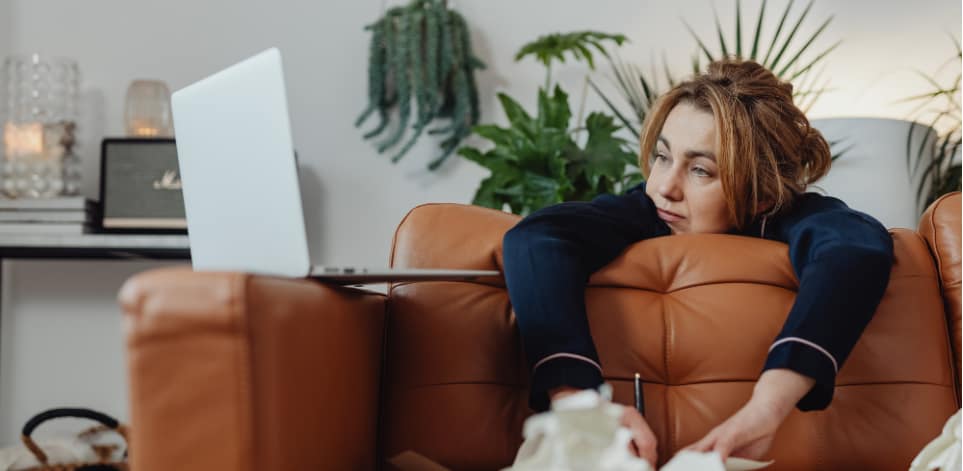 A woman slumped on a sofa with her laptop open, looking mentally exhausted while online shopping, surrounded by a calm home setting.